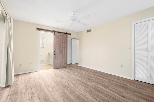 unfurnished bedroom featuring ceiling fan, ensuite bathroom, light hardwood / wood-style flooring, and a barn door