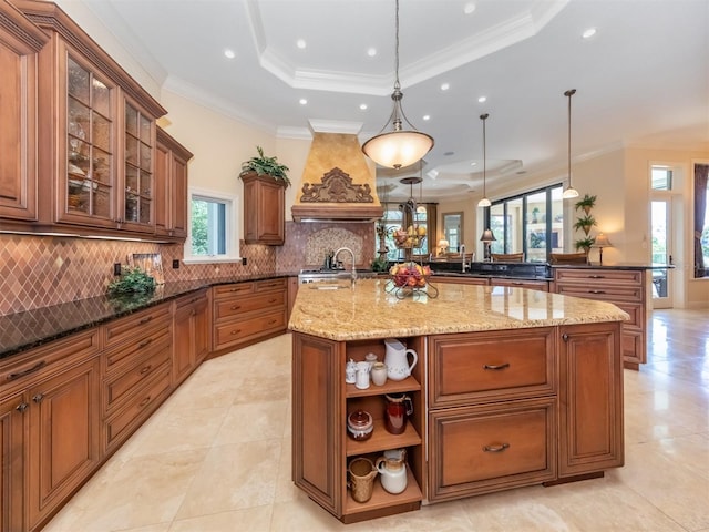 kitchen featuring light stone countertops, premium range hood, a raised ceiling, hanging light fixtures, and an island with sink