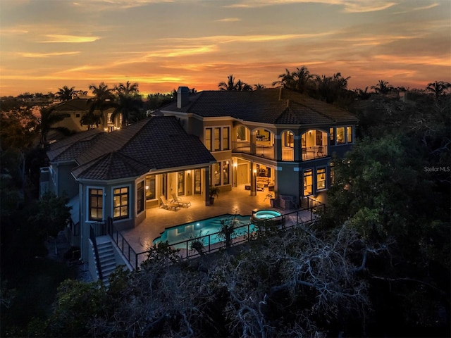 back house at dusk featuring a balcony, a patio area, and a pool with hot tub