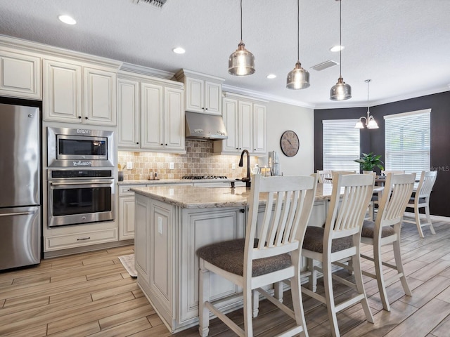 kitchen featuring appliances with stainless steel finishes, a center island with sink, light stone counters, and hanging light fixtures