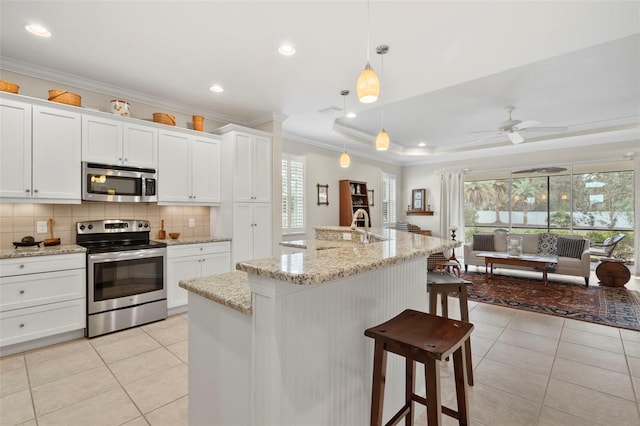 kitchen with pendant lighting, a raised ceiling, an island with sink, white cabinetry, and stainless steel appliances