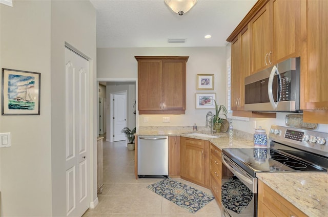 kitchen with brown cabinetry, light stone countertops, stainless steel appliances, a sink, and light tile patterned flooring