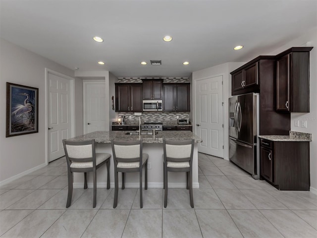 kitchen featuring light stone counters, an island with sink, appliances with stainless steel finishes, and dark brown cabinetry