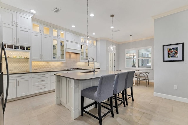 kitchen with white cabinetry, premium range hood, hanging light fixtures, ornamental molding, and a kitchen island with sink