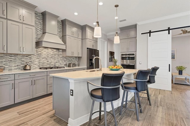 kitchen with sink, stainless steel appliances, custom range hood, a center island with sink, and a barn door