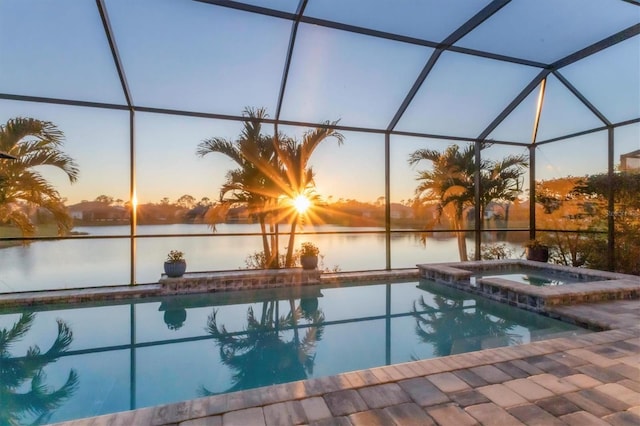 pool at dusk featuring an in ground hot tub, a water view, a patio area, and glass enclosure