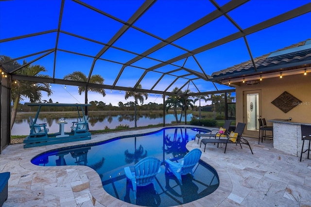 pool at dusk with a patio area, glass enclosure, a water view, and an outdoor bar