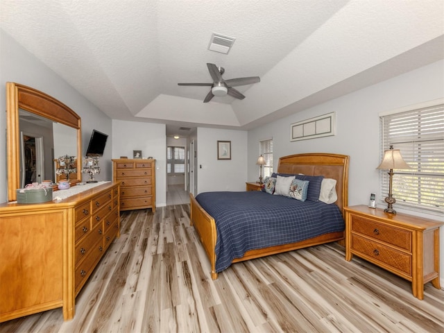 bedroom with light wood-type flooring, ensuite bath, a textured ceiling, a tray ceiling, and ceiling fan