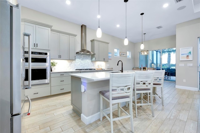 kitchen with a breakfast bar area, stainless steel appliances, gray cabinetry, a kitchen island with sink, and wall chimney exhaust hood