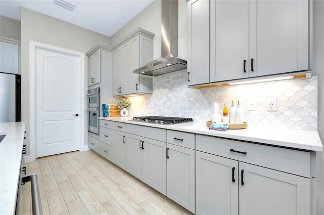 kitchen featuring appliances with stainless steel finishes, wall chimney exhaust hood, decorative backsplash, light hardwood / wood-style floors, and gray cabinets