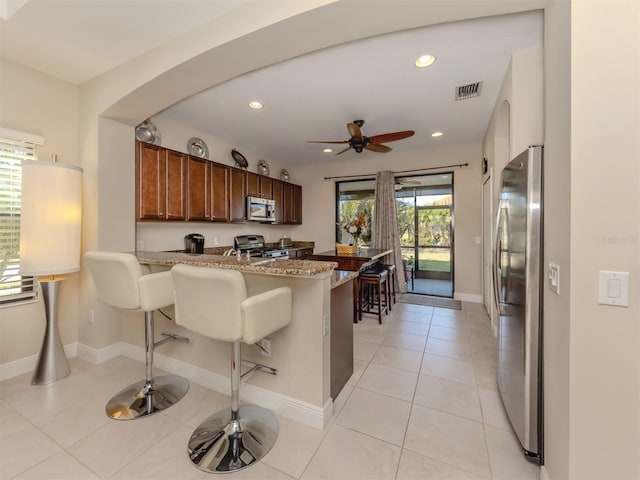 kitchen with appliances with stainless steel finishes, kitchen peninsula, light stone countertops, a breakfast bar, and light tile patterned flooring