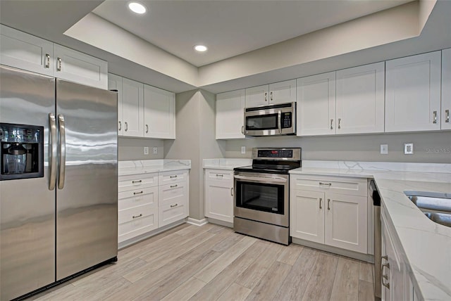 kitchen featuring white cabinetry, a tray ceiling, stainless steel appliances, light stone countertops, and light hardwood / wood-style floors