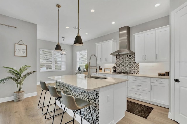 kitchen featuring pendant lighting, a kitchen island with sink, wall chimney range hood, sink, and white cabinetry