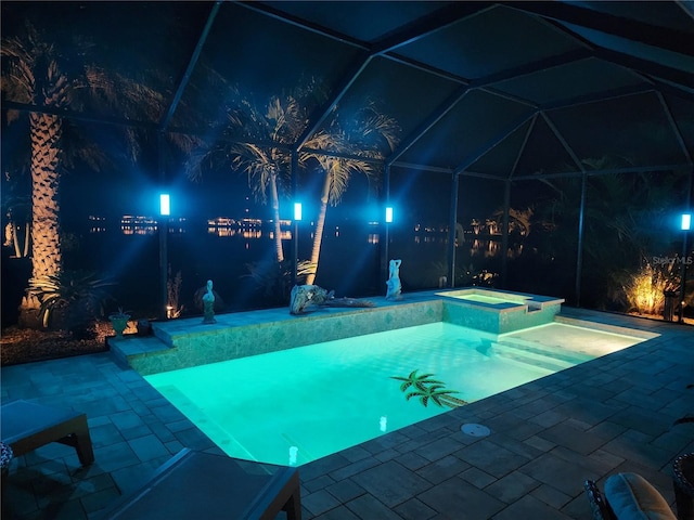 pool at night featuring a patio area, a water view, glass enclosure, and an in ground hot tub