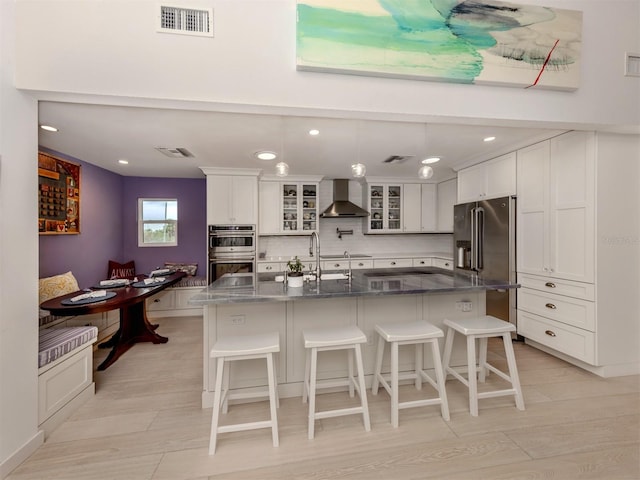 kitchen with backsplash, a large island with sink, wall chimney range hood, white cabinetry, and stainless steel appliances