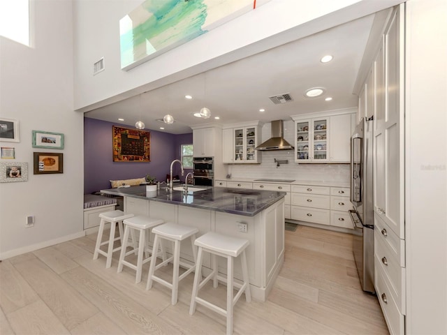 kitchen with a kitchen bar, a center island with sink, backsplash, wall chimney exhaust hood, and white cabinets