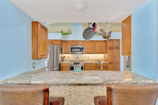kitchen featuring brown cabinets, light stone countertops, appliances with stainless steel finishes, a peninsula, and a kitchen bar