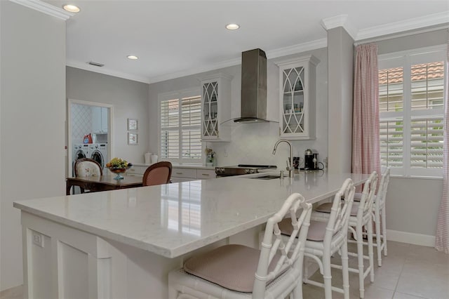 kitchen featuring wall chimney exhaust hood, sink, independent washer and dryer, light tile patterned flooring, and a breakfast bar area