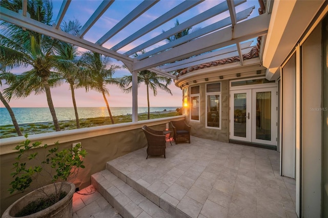 patio terrace at dusk with a beach view, french doors, a water view, and a pergola