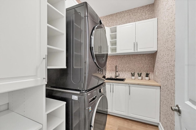 clothes washing area featuring light wood finished floors, stacked washer / drying machine, cabinet space, a sink, and wallpapered walls