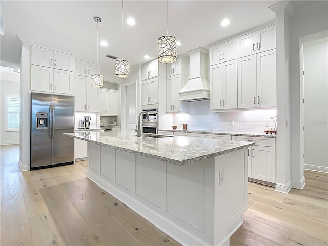 kitchen featuring white cabinets, custom range hood, an island with sink, and appliances with stainless steel finishes