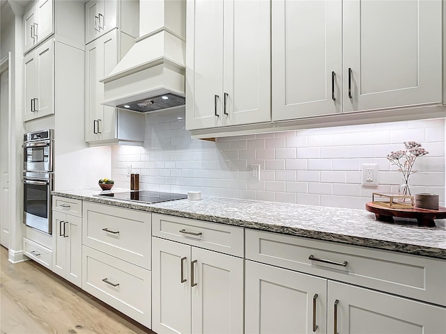 kitchen with custom exhaust hood, white cabinets, light stone counters, and double oven