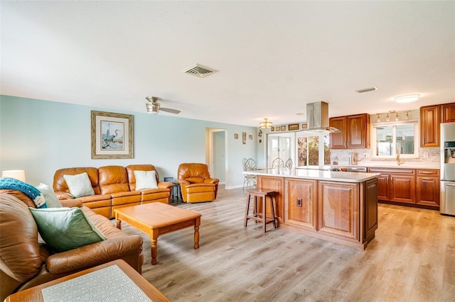 kitchen with island exhaust hood, tasteful backsplash, light hardwood / wood-style floors, stainless steel fridge, and a center island with sink