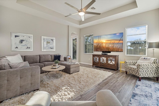 living area with arched walkways, ceiling fan, wood finished floors, baseboards, and a tray ceiling