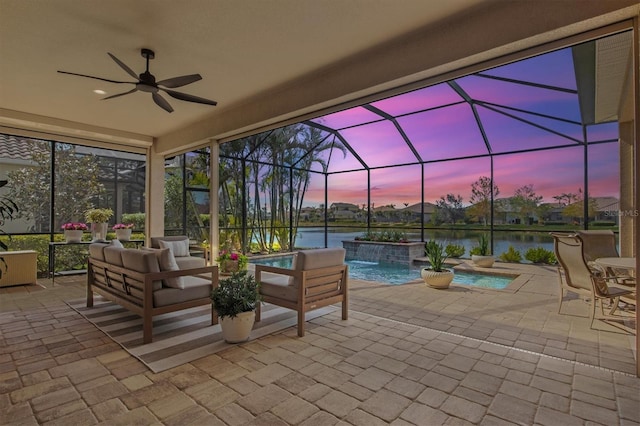 patio terrace at dusk with a lanai, ceiling fan, a water view, an outdoor living space, and pool water feature