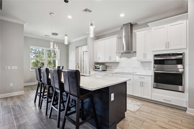 kitchen featuring pendant lighting, an island with sink, white cabinetry, stainless steel double oven, and wall chimney exhaust hood