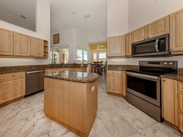 kitchen with dark stone countertops, stainless steel appliances, a center island, a high ceiling, and light brown cabinetry