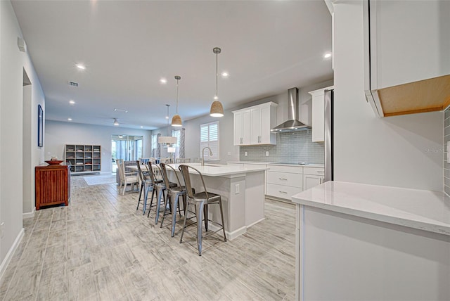 kitchen with a breakfast bar, decorative light fixtures, white cabinets, wall chimney range hood, and a center island with sink