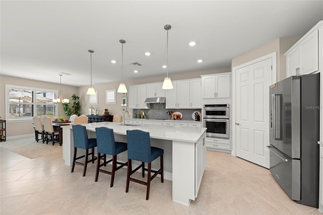 kitchen featuring white cabinetry, decorative light fixtures, an island with sink, stainless steel appliances, and backsplash