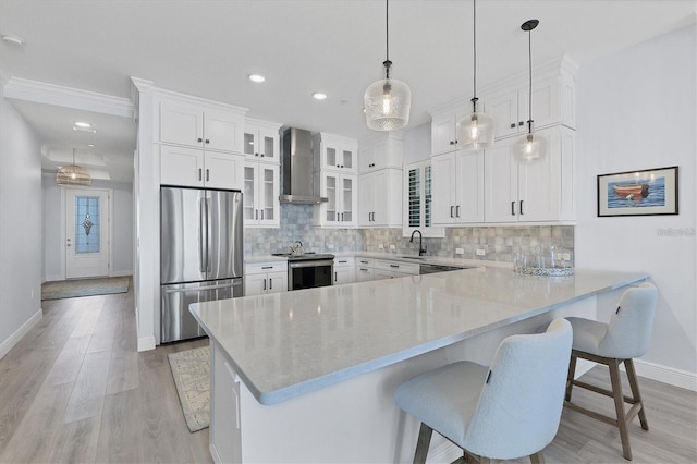 kitchen featuring a breakfast bar area, stainless steel appliances, white cabinets, decorative light fixtures, and wall chimney exhaust hood