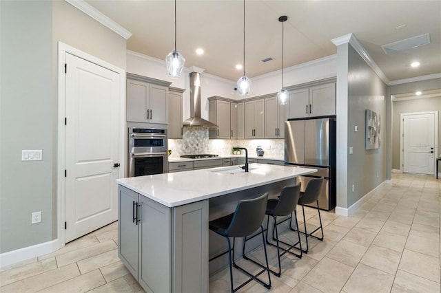 kitchen with wall chimney exhaust hood, appliances with stainless steel finishes, gray cabinets, a kitchen island with sink, and backsplash