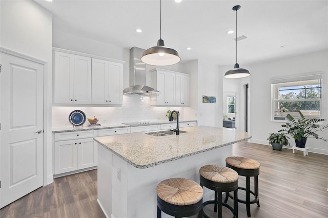 kitchen featuring sink, decorative light fixtures, an island with sink, wall chimney range hood, and white cabinets