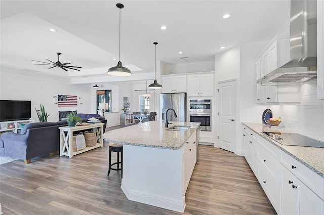 kitchen with sink, white cabinetry, an island with sink, stainless steel appliances, and wall chimney range hood