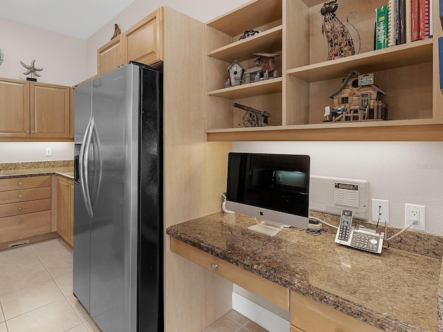 kitchen featuring stainless steel refrigerator with ice dispenser, built in desk, light tile patterned floors, and dark stone countertops