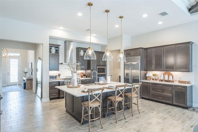 kitchen featuring pendant lighting, an island with sink, a kitchen breakfast bar, built in appliances, and wall chimney exhaust hood