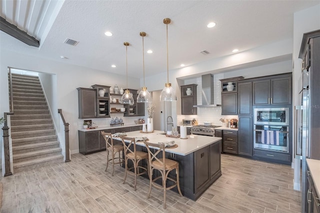 kitchen featuring wall chimney exhaust hood, stainless steel appliances, a kitchen island with sink, and dark brown cabinets