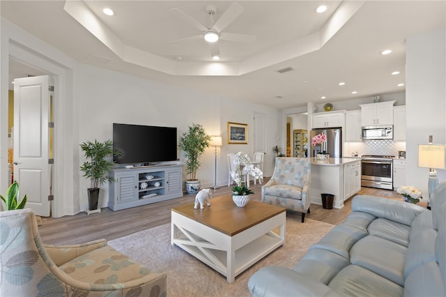 living room with a raised ceiling, ceiling fan, and light hardwood / wood-style flooring