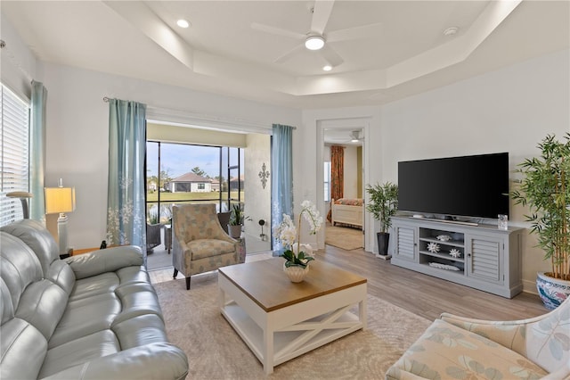 living room with a raised ceiling, ceiling fan, and light wood-type flooring