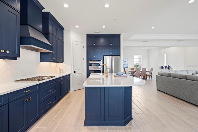 kitchen featuring blue cabinets, stainless steel appliances, premium range hood, and a center island with sink