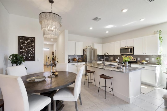 kitchen featuring appliances with stainless steel finishes, a kitchen island with sink, hanging light fixtures, white cabinetry, and dark stone counters