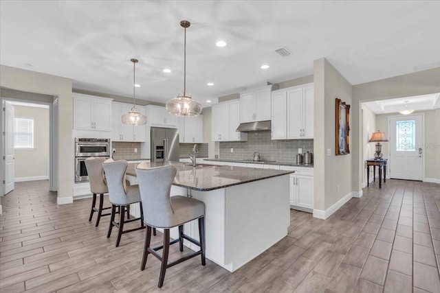 kitchen featuring decorative light fixtures, a center island with sink, appliances with stainless steel finishes, dark stone counters, and white cabinets