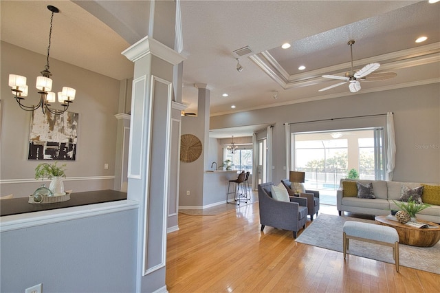 living room featuring decorative columns, ornamental molding, a tray ceiling, light hardwood / wood-style floors, and ceiling fan with notable chandelier