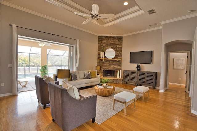 living room featuring light hardwood / wood-style flooring, ceiling fan, ornamental molding, a stone fireplace, and a raised ceiling