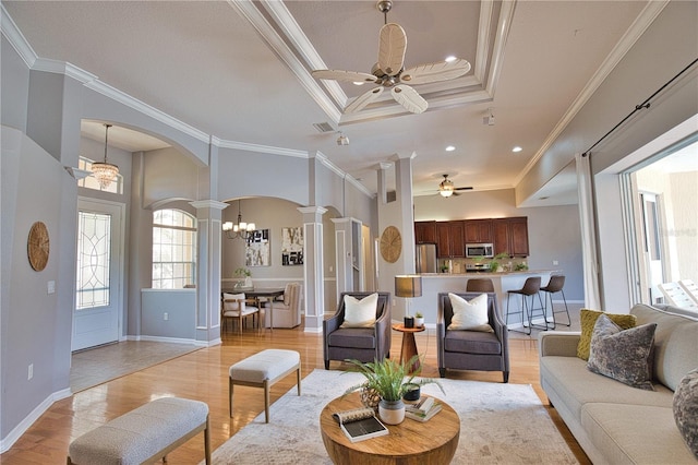 living room featuring ornate columns, ceiling fan with notable chandelier, ornamental molding, a tray ceiling, and light wood-type flooring