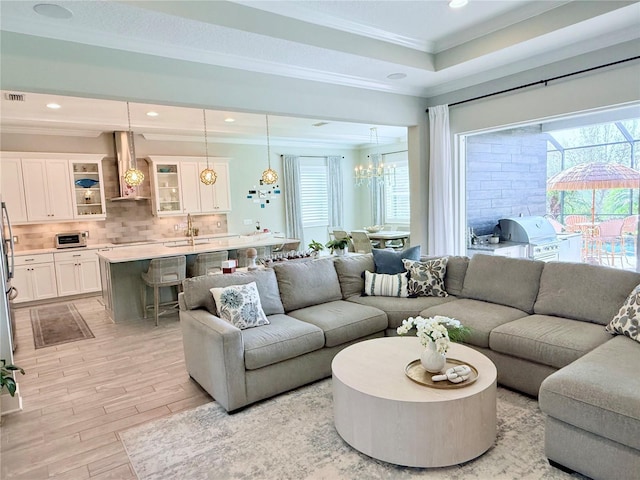 living room with sink, a notable chandelier, a tray ceiling, crown molding, and light hardwood / wood-style flooring