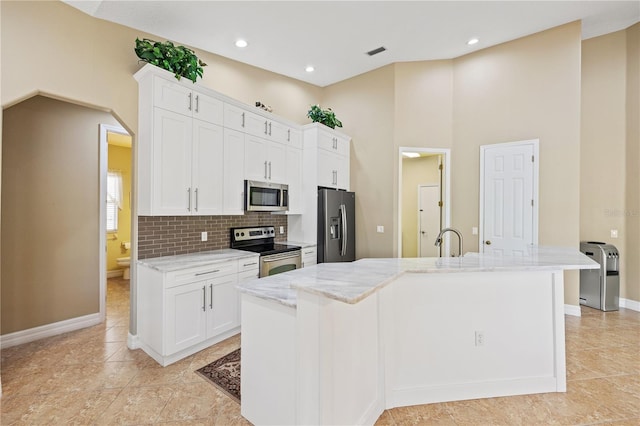 kitchen with light stone counters, appliances with stainless steel finishes, a kitchen island with sink, decorative backsplash, and white cabinets